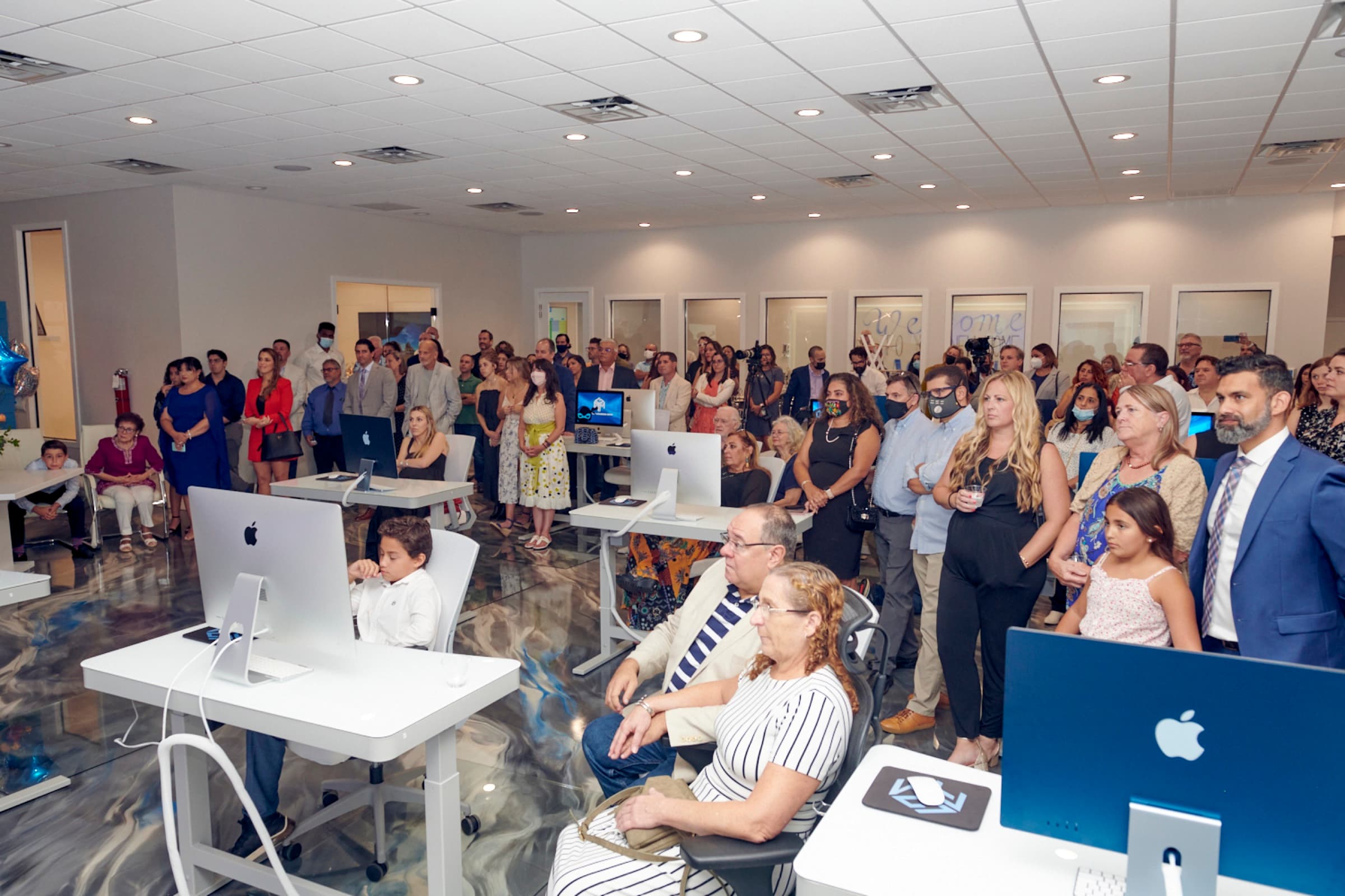 A packed grand-opening crowd standing among the white desks and iMacs of the training floor